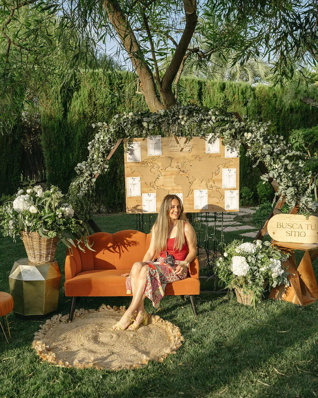 Rincón de seating con un mapa del mundo de madera, arco de eucalipto y un sofá naranja bajo los árboles