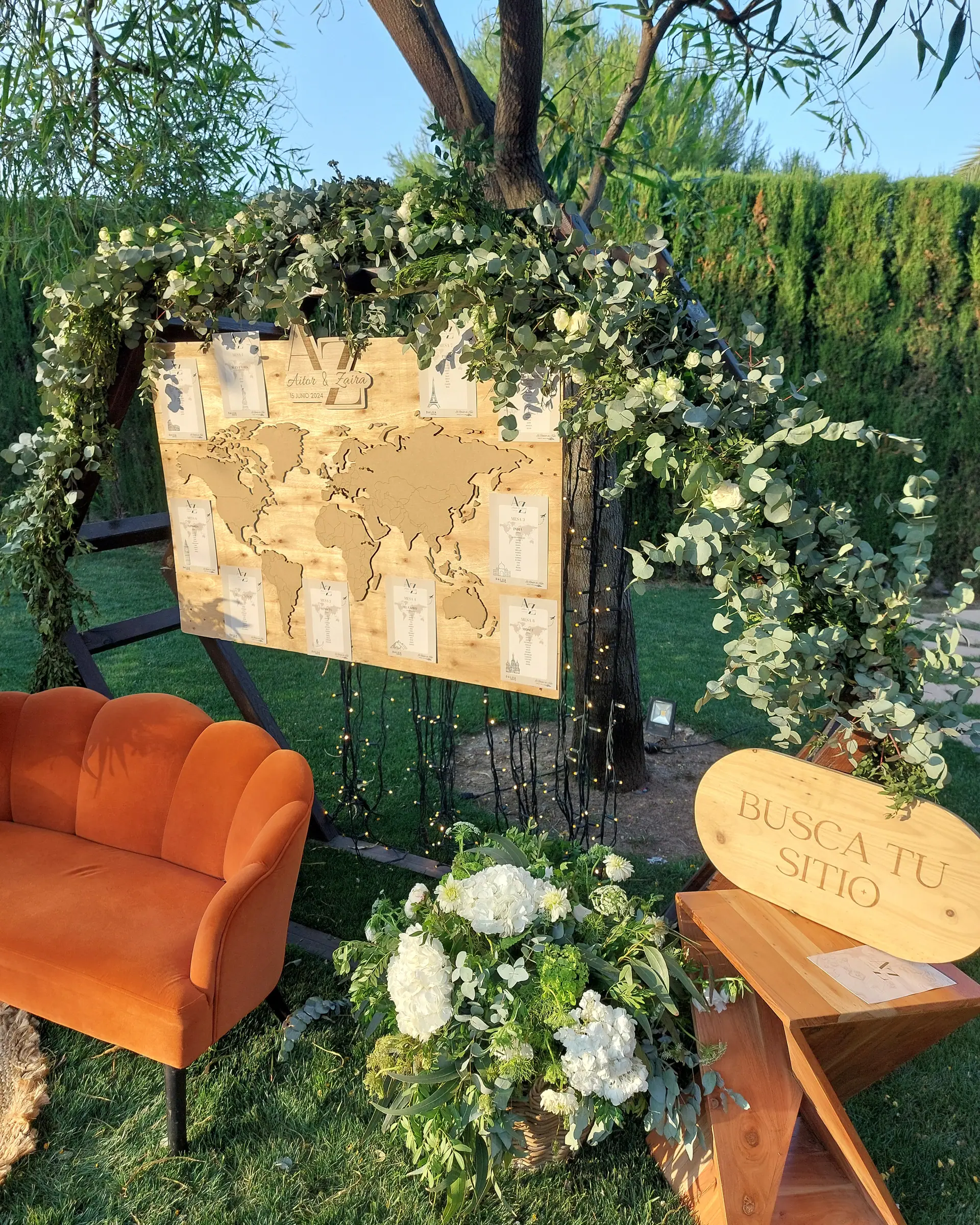 Seating plan con el mapa del mundo grabado en madera, colgado bajo un arco de eucalipto, con un sofá de terciopelo y el cartel «Busca tu sitio»